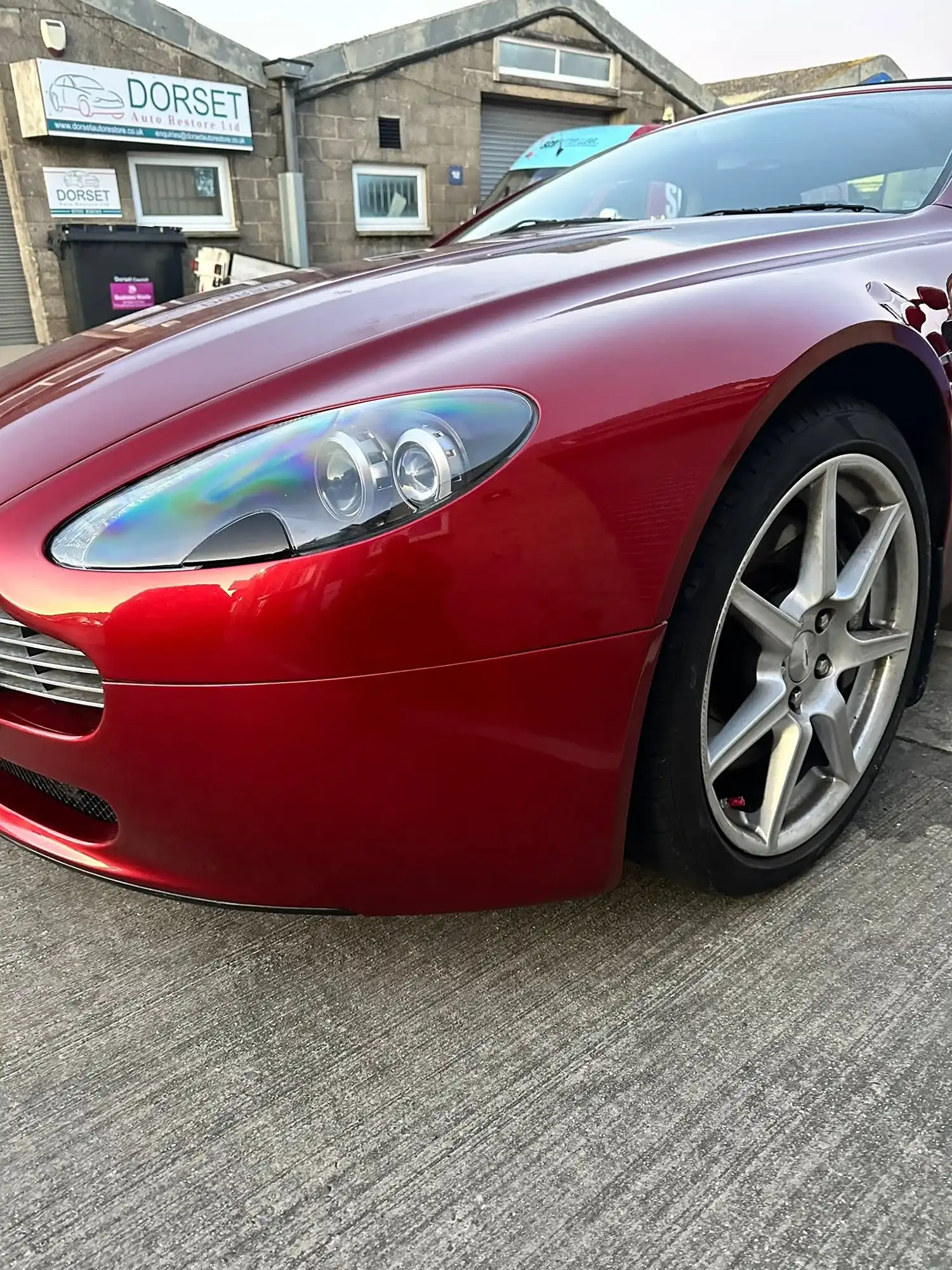 Close-up of front left side of a shiny red sports car parked on concrete near a stone building.