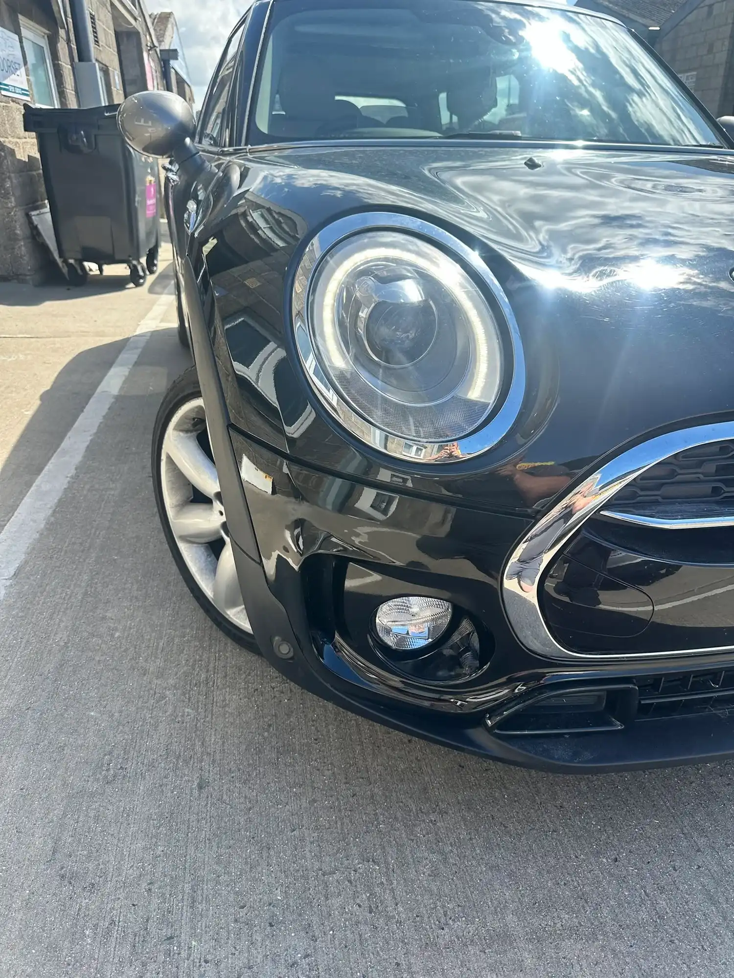 Close-up of a black car's front left headlight and wheel parked on a concrete road.
