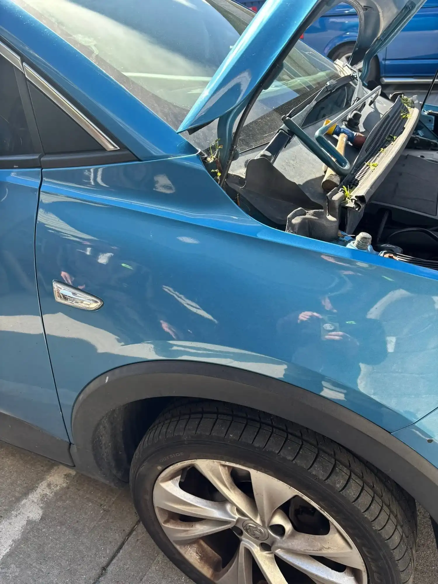 Blue car with open hood showing tools and small plants growing inside near front wheel.