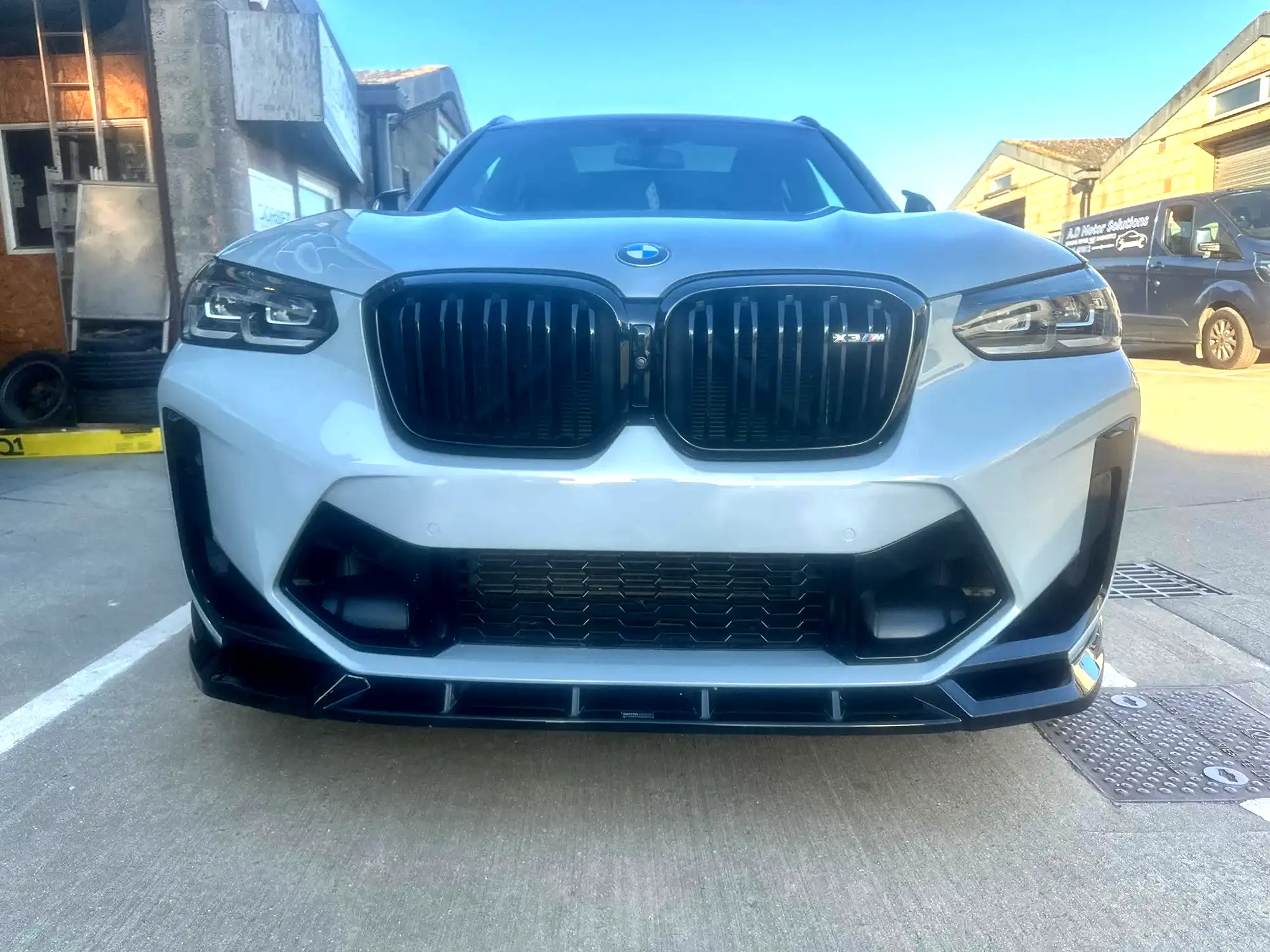 Front view of a white BMW X3 M parked on concrete near buildings and a black van.