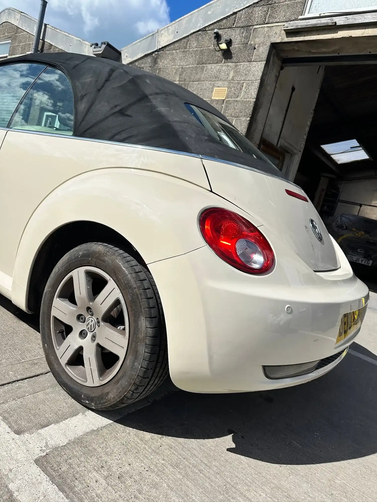 Rear side view of a cream Volkswagen Beetle convertible parked near a garage.