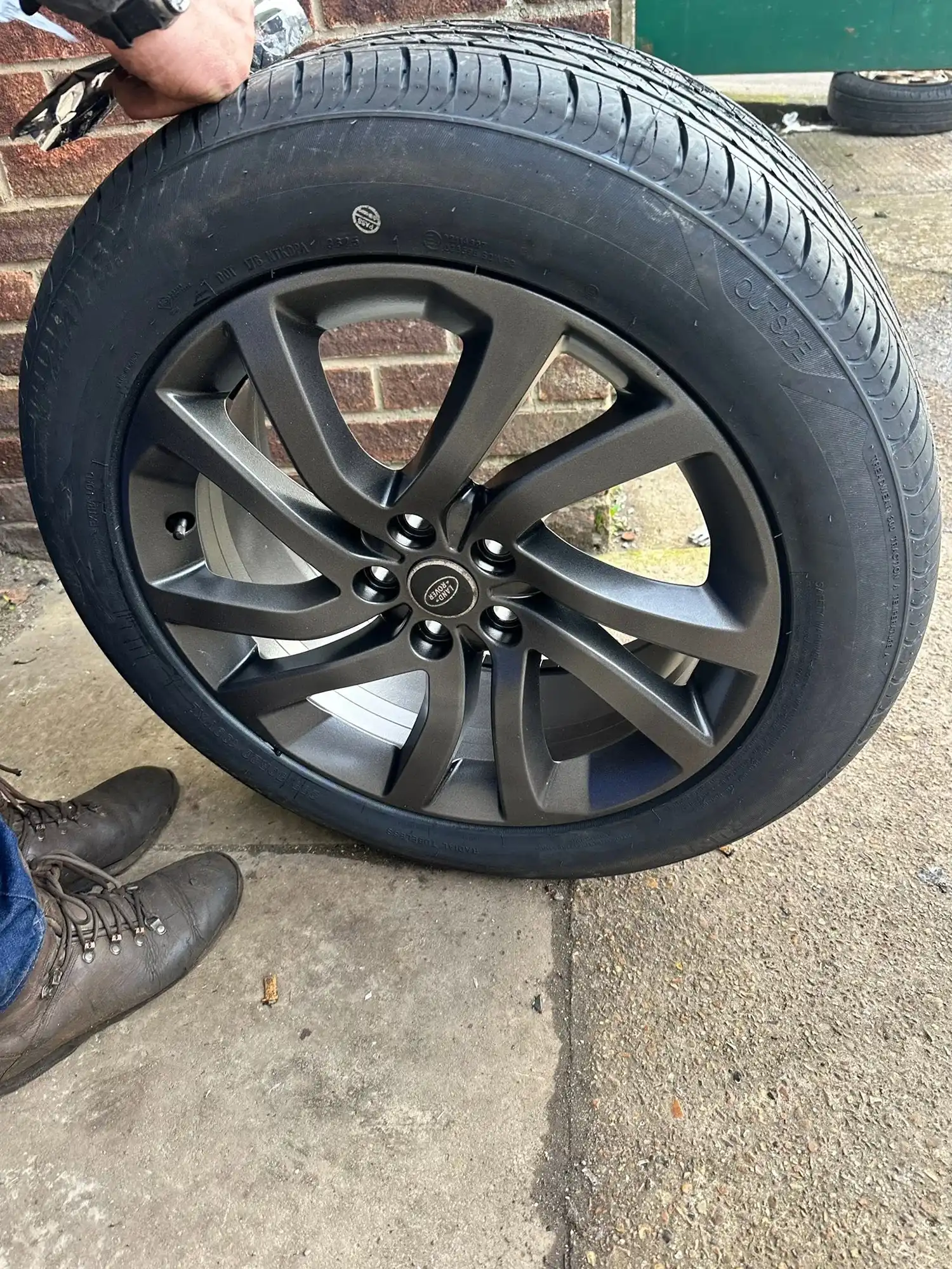 Close-up of a Land Rover wheel and tire on concrete with a person's hand and boots visible.