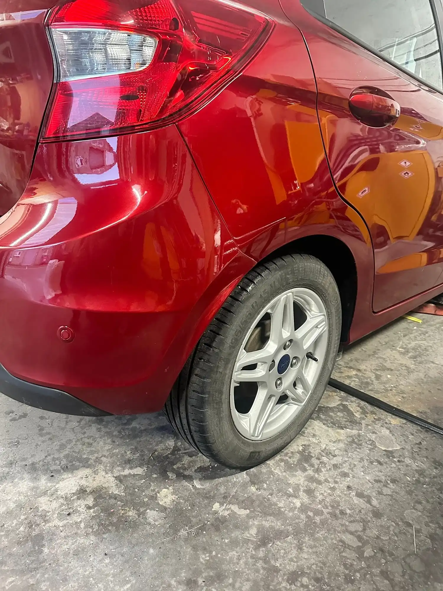 Close-up of a red car's rear side, showing the taillight, door, and rear tire on a concrete floor.